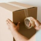 Crop anonymous male using tape to seal packed cardboard box against white wall while moving into new place